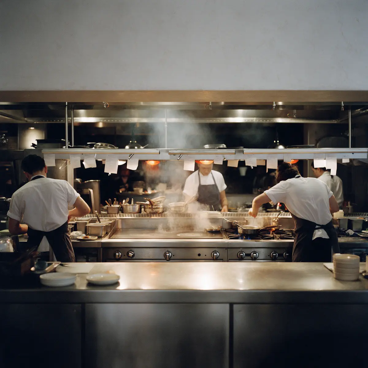 A restaurant kitchen mid-service: cooks at the line, steam, order tickets on the rail.